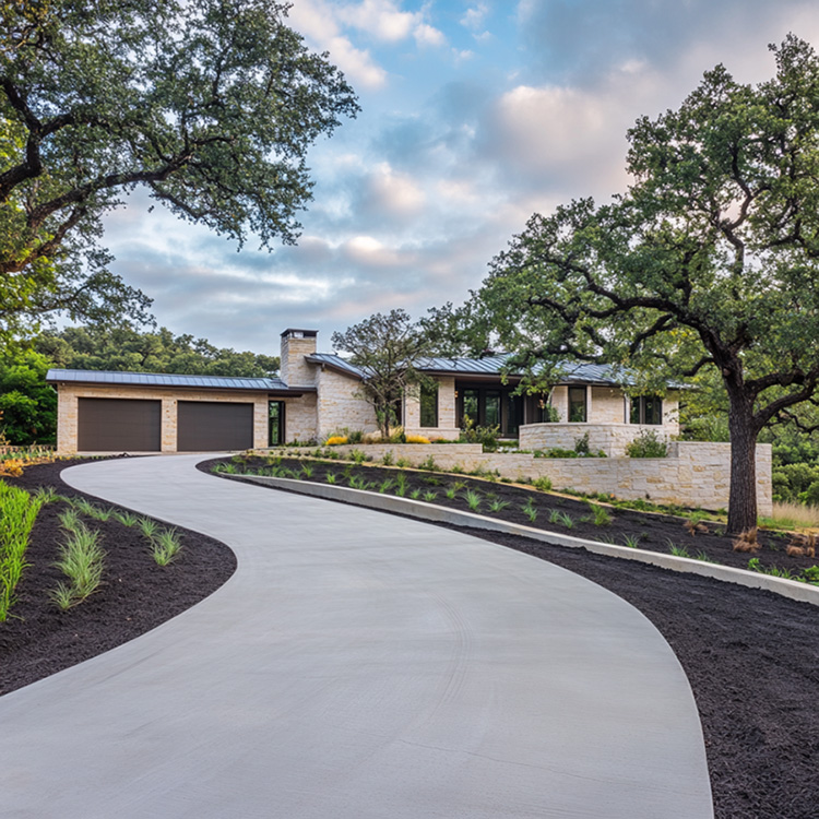 a newly finished concrete driveway winding uphill toward a large, modern ranch-style home in Abilene, Texas. The driveway is expansive and multi-directional, curving gracefully through a landscaped front yard with mature oak trees, dark mulch beds, and exposed limestone accents.